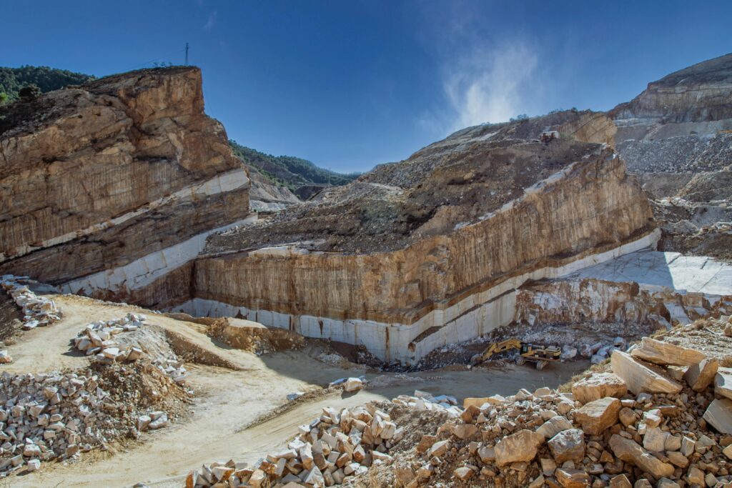 Aerial shot of a quarry displaying geological layers and an excavator amidst rocky terrain under a clear blue sky.